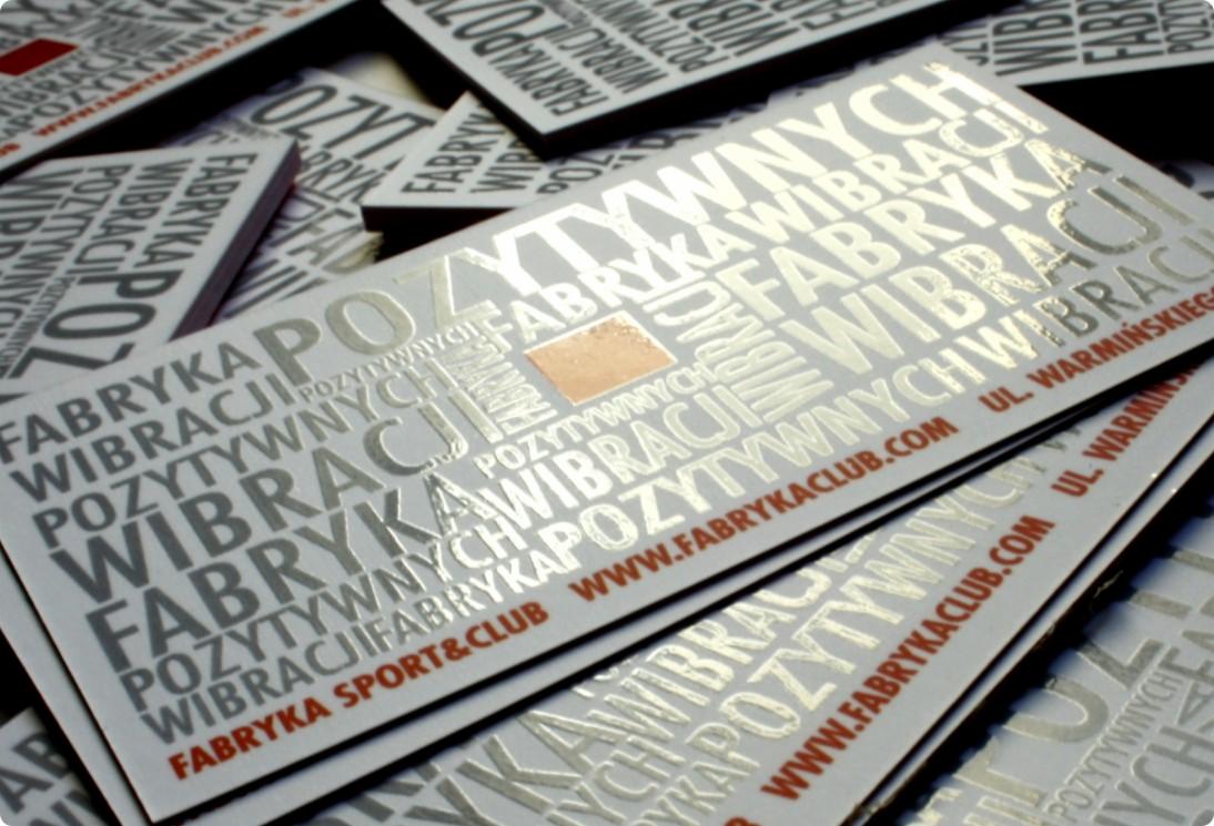 Stos wizytówek z srebrnymi, tłoczonymi napisami 'Fabryka Pozytywnych Wibracji' i adresem strony internetowej fabrykaclub.com oraz adresem.
