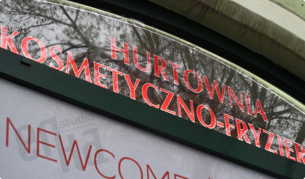 Zewnętrzny szyld z czerwonym napisem 'KOSMETYCZNO-FRYZJER HURTOWNIA' na przezroczystym tle, widoczne odbicie drzew, poniżej napis 'NEWCOM', ujęcie pod kątem.