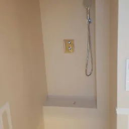 Wykończona kabina prysznicowa walk-in z deszczownicą, słuchawką prysznicową na ścianie, półką i dwoma chromowanymi pokrętłami regulacji temperatury.