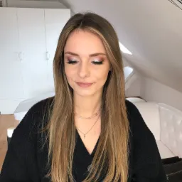 Portret kobiety z subtelnym, błyszczącym makijażem oczu i delikatnym makijażem ust w Opace. Widoczne długie, proste włosy w odcieniach blondu i brązu oraz czarny sweter. W tle białe meble i skośny...