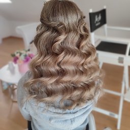 Elżbieta Lis Make-Up - Upięcie z fal, korona na głowie, widok z tyłu. Jasne, kręcone włosy z warkoczami, stylizacja ślubna w salonie. W tle krzesło reżyserskie i kwiaty.