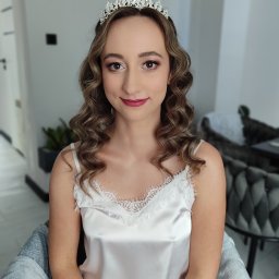 Elżbieta Lis Make-Up - Portret kobiety z delikatnym upięciem, koroną i subtelnym makijażem w odcieniach brązu i różu, siedzącej na krześle w jasnym wnętrzu.
