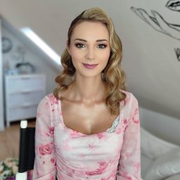 Elżbieta Lis Make-Up - Portret kobiety z profesjonalnym makijażem i upiętymi blond włosami w stylu retro, w bluzce w różowe róże, na jasnym tle z rysunkiem.