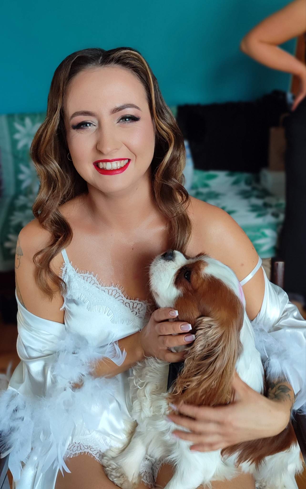 Uśmiechnięta kobieta z czerwonymi ustami, w białym szlafroku z piórami, trzymająca psa rasy Cavalier King Charles Spaniel. Widoczne delikatne fale na włosach.