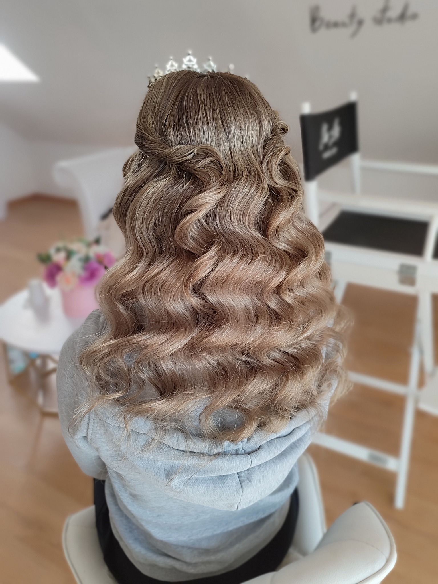 Upięcie z fal, korona na głowie, widok z tyłu. Jasne, kręcone włosy z warkoczami, stylizacja ślubna w salonie. W tle krzesło reżyserskie i kwiaty.