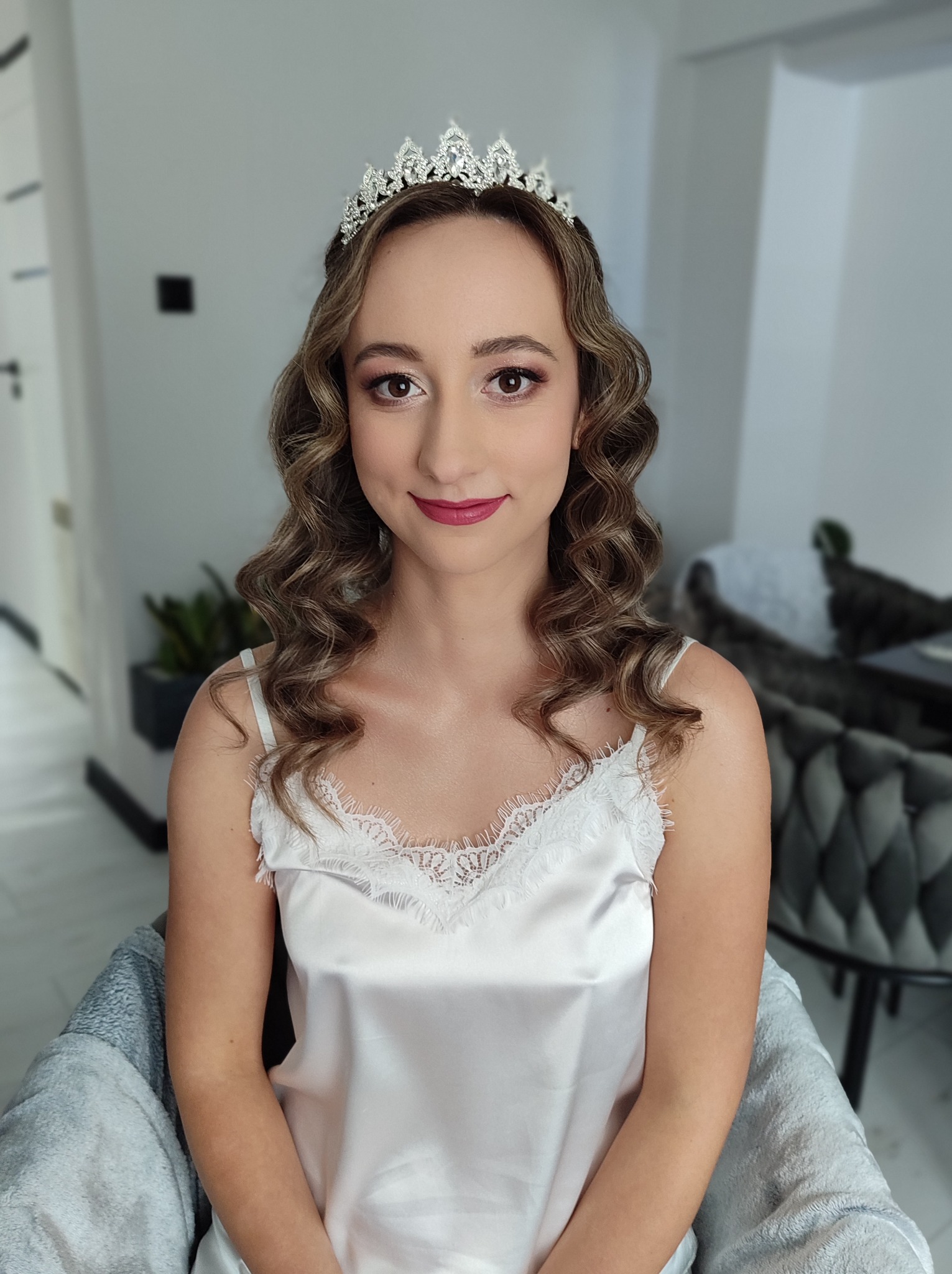 Portret kobiety z delikatnym upięciem, koroną i subtelnym makijażem w odcieniach brązu i różu, siedzącej na krześle w jasnym wnętrzu.