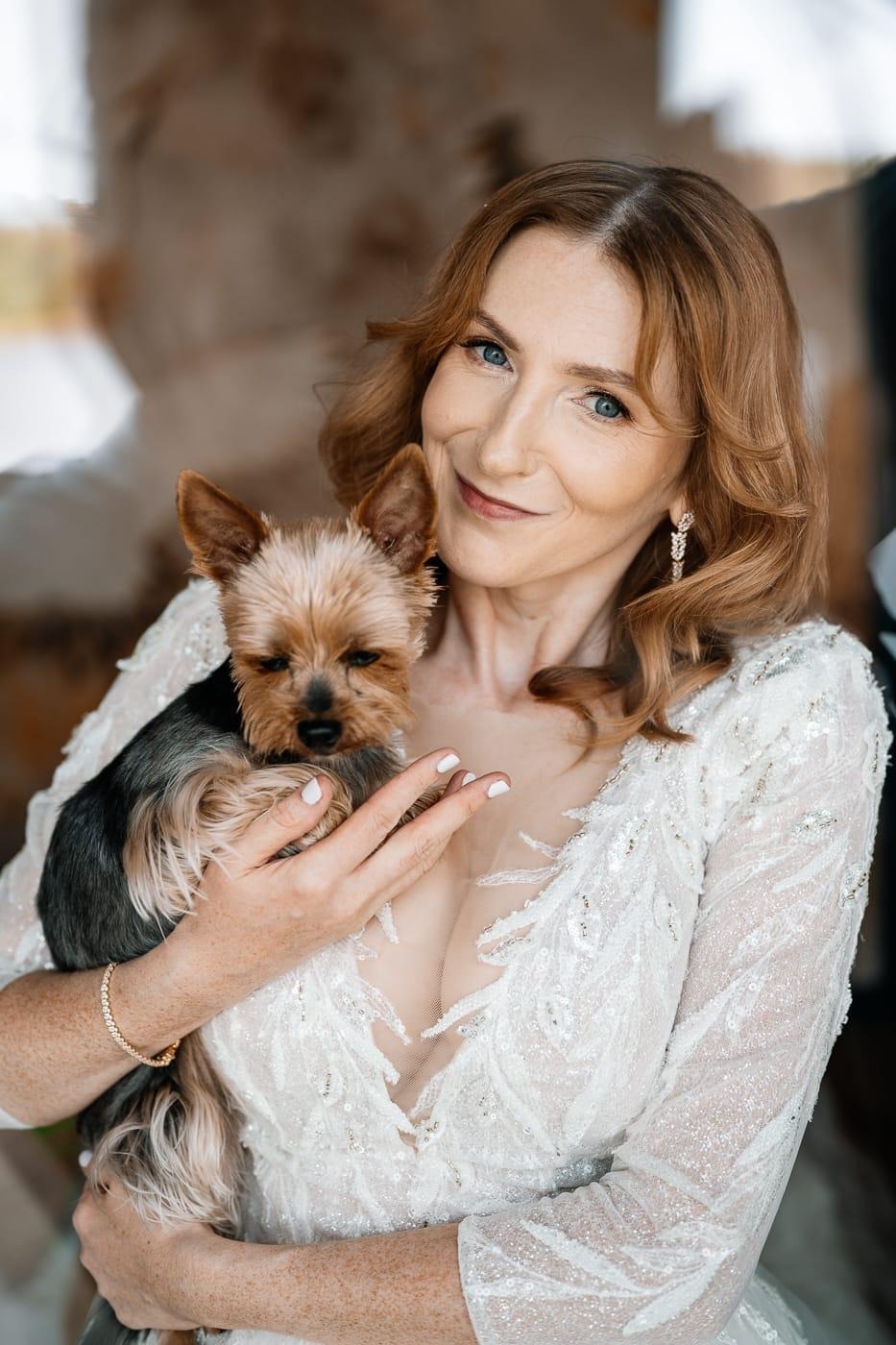 Uśmiechnięta kobieta w białej sukni trzyma Yorka. Delikatny makijaż, upięte włosy i błyszcząca biżuteria. Portret w ciepłych tonacjach.