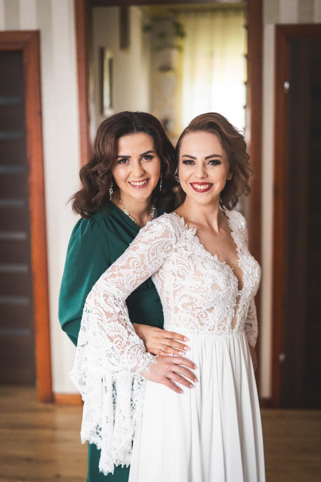 Uśmiechnięta panna młoda w koronkowej sukni ślubnej pozuje z druhną w szmaragdowej sukni, prezentując upięcie i makijaż ślubny w jasnym wnętrzu.