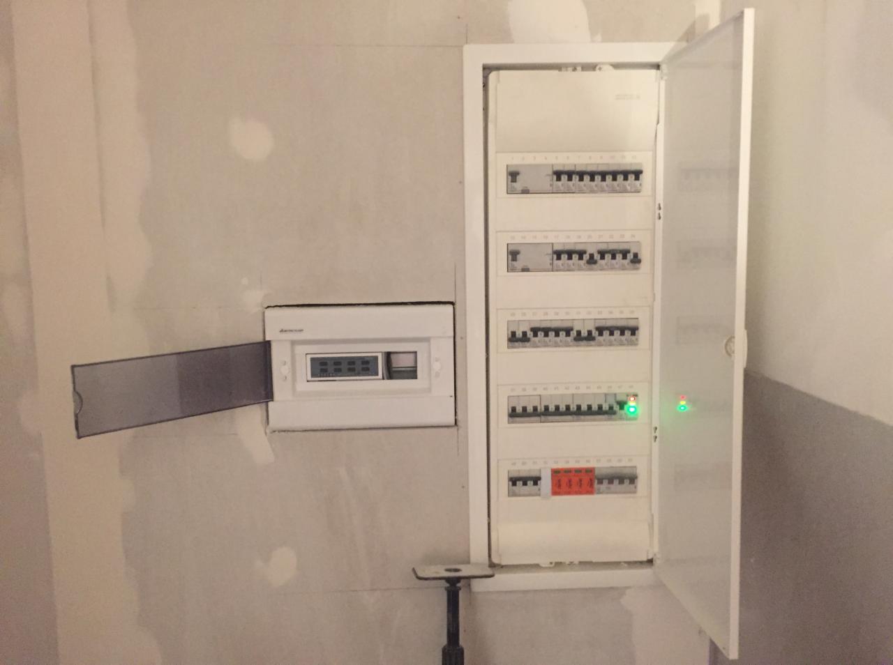 Dwie białe skrzynki rozdzielcze na surowej ścianie, jedna z otwartą klapką i druga z otwartymi drzwiczkami, widoczne zabezpieczenia i włączniki, zielona dioda sygnalizacyjna na jednym z paneli.