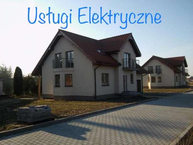 Dwa nowe domy jednorodzinne z czerwonymi dachami i balkonami, widoczne z poziomu brukowanej drogi w słoneczny dzień. Na pierwszym planie stos kostki brukowej.