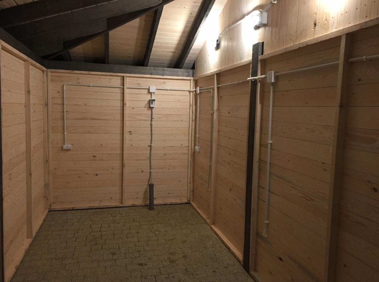 Wnętrze pomieszczenia wykończonego boazerią z widocznymi przewodami i puszkami elektrycznymi, oświetleniem sufitowym i podłogą z kostki brukowej.