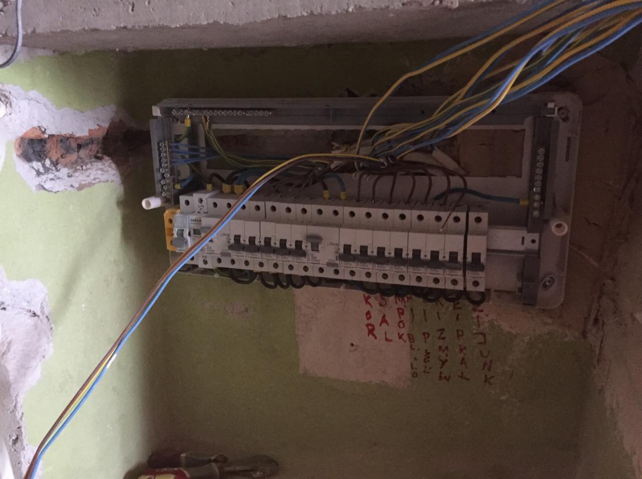 Rozdzielnica elektryczna z podłączonymi przewodami w surowym stanie, widoczne ślady po kuciu w ścianie i napisy na ścianie wykonane pisakiem.