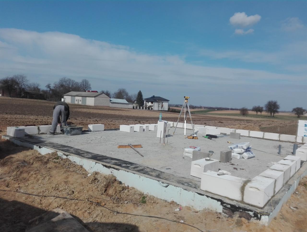 Budowa fundamentów domu z bloczków betonowych; widoczny pracownik, poziomica laserowa, bloczki, worki z cementem i otwarty krajobraz w tle.