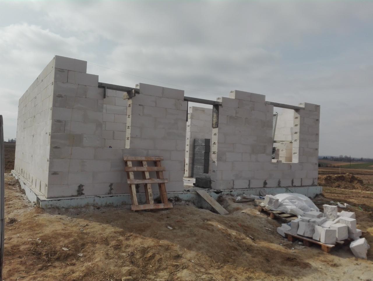 Wznoszone ściany parterowego domu z białych bloczków, widoczne zbrojenie nad otworami okiennymi, drewniana paleta i porozrzucane bloczki na ziemi.