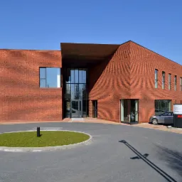 Nowoczesny budynek biurowy z czerwonej cegły w Katowicach, z przeszklonym wejściem i okrągłym klombem z trawą przed budynkiem. Na parkingu zaparkowane samochody.