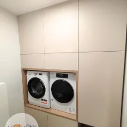 Jasna zabudowa meblowa w łazience z pralką i suszarką. Minimalistyczny design, gładkie fronty, drewniane akcenty. Funkcjonalna przestrzeń.