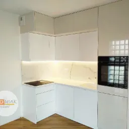 Biała, nowoczesna kuchnia z marmurowym blatem i oświetleniem LED. Widoczna płyta indukcyjna i piekarnik w zabudowie. Minimalistyczny design z połyskującymi frontami.