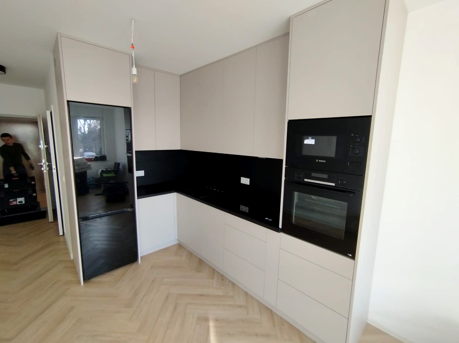 Jasna kuchnia z czarnym panelem i sprzętem Bosch, drewniana podłoga w jodełkę. Widoczny pracownik w tle.