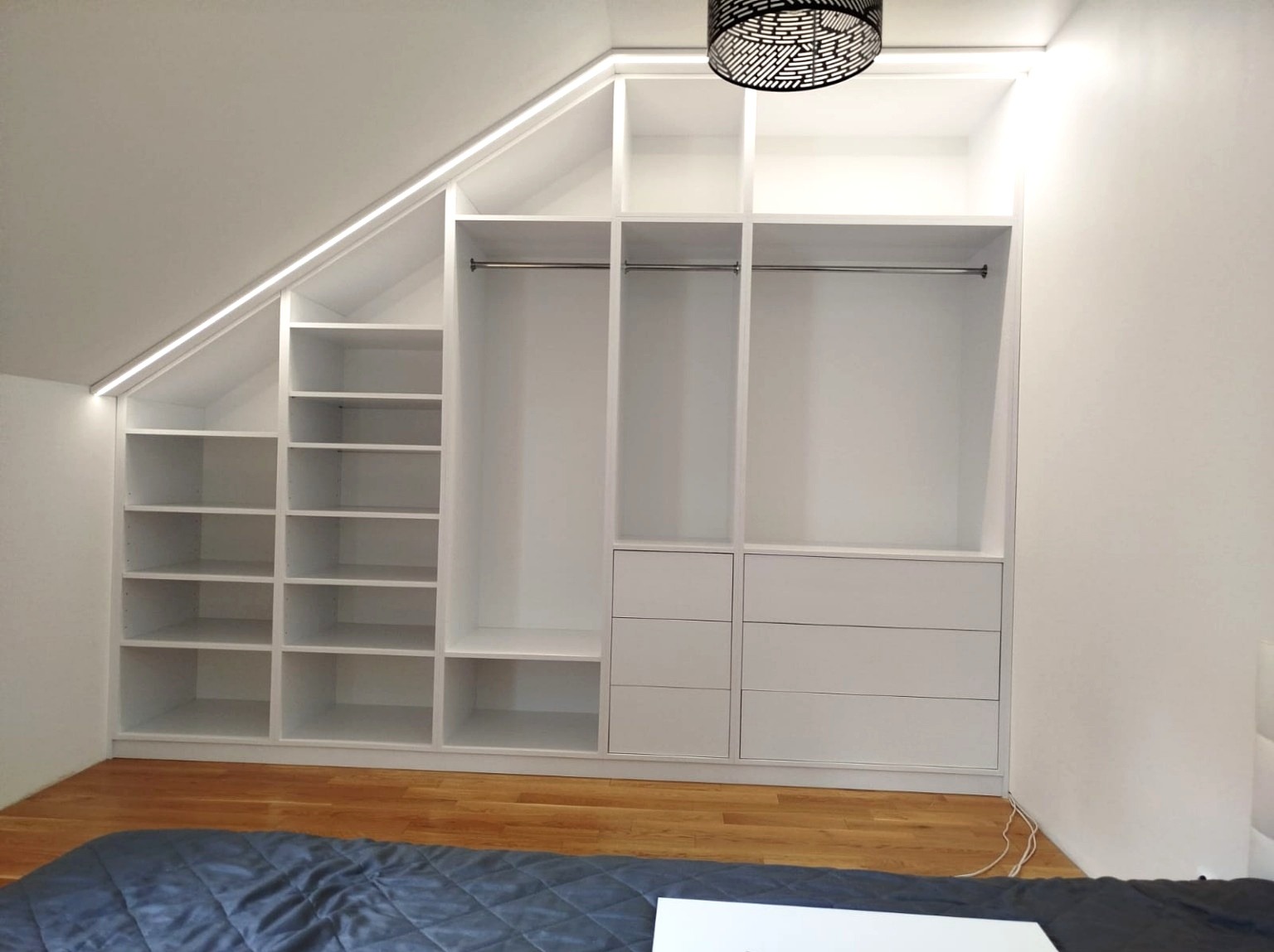Biała szafa wnękowa z półkami i drążkami, dopasowana do skosu poddasza, oświetlona listwą LED. Widoczny fragment łóżka i parkietu.