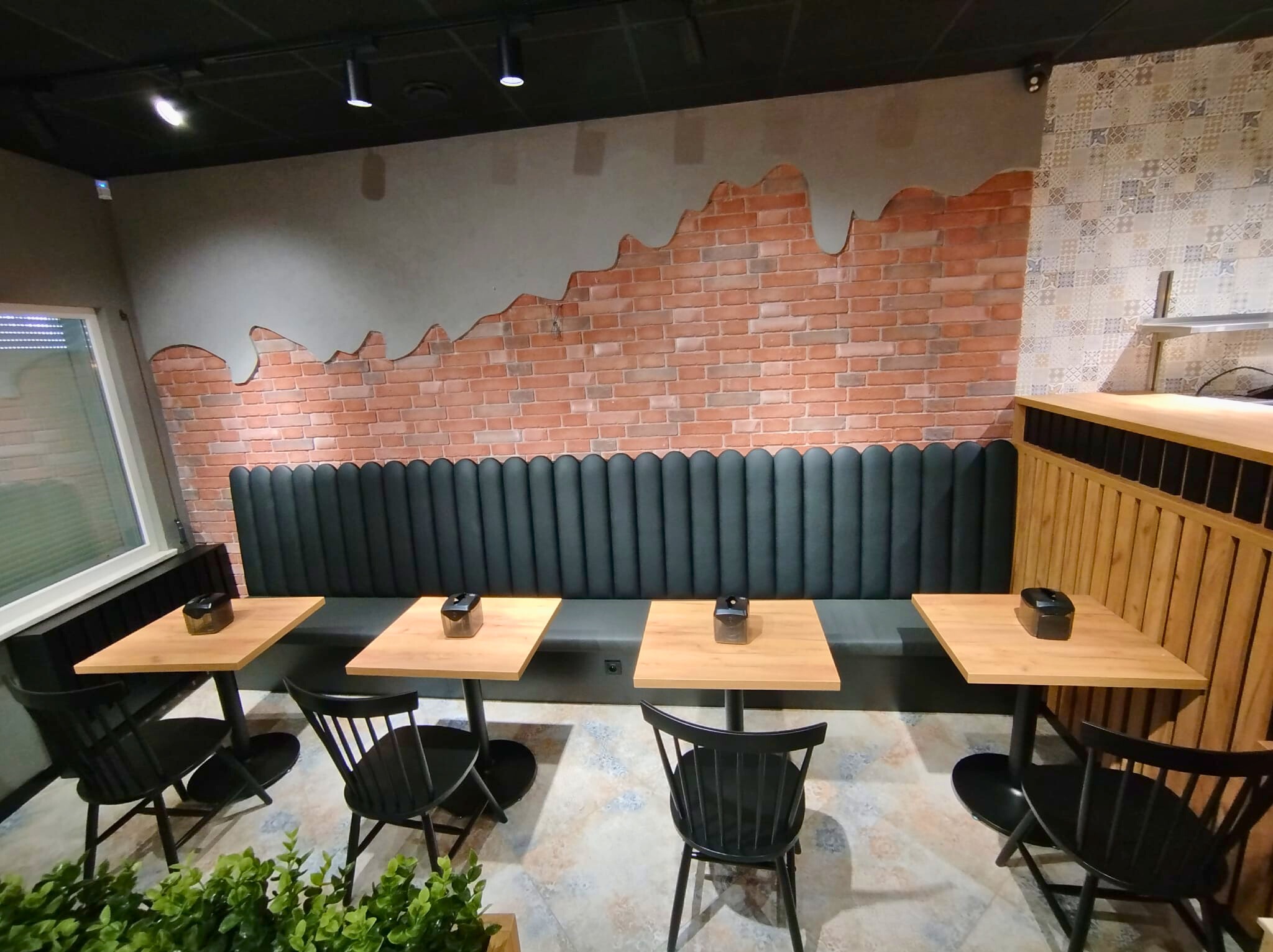 Wnętrze lokalu z tapicerowanym siedziskiem wzdłuż ceglanej ściany, drewnianymi stolikami na czarnych nogach i czarnymi krzesłami. Uwagę zwraca dekoracja ściany.