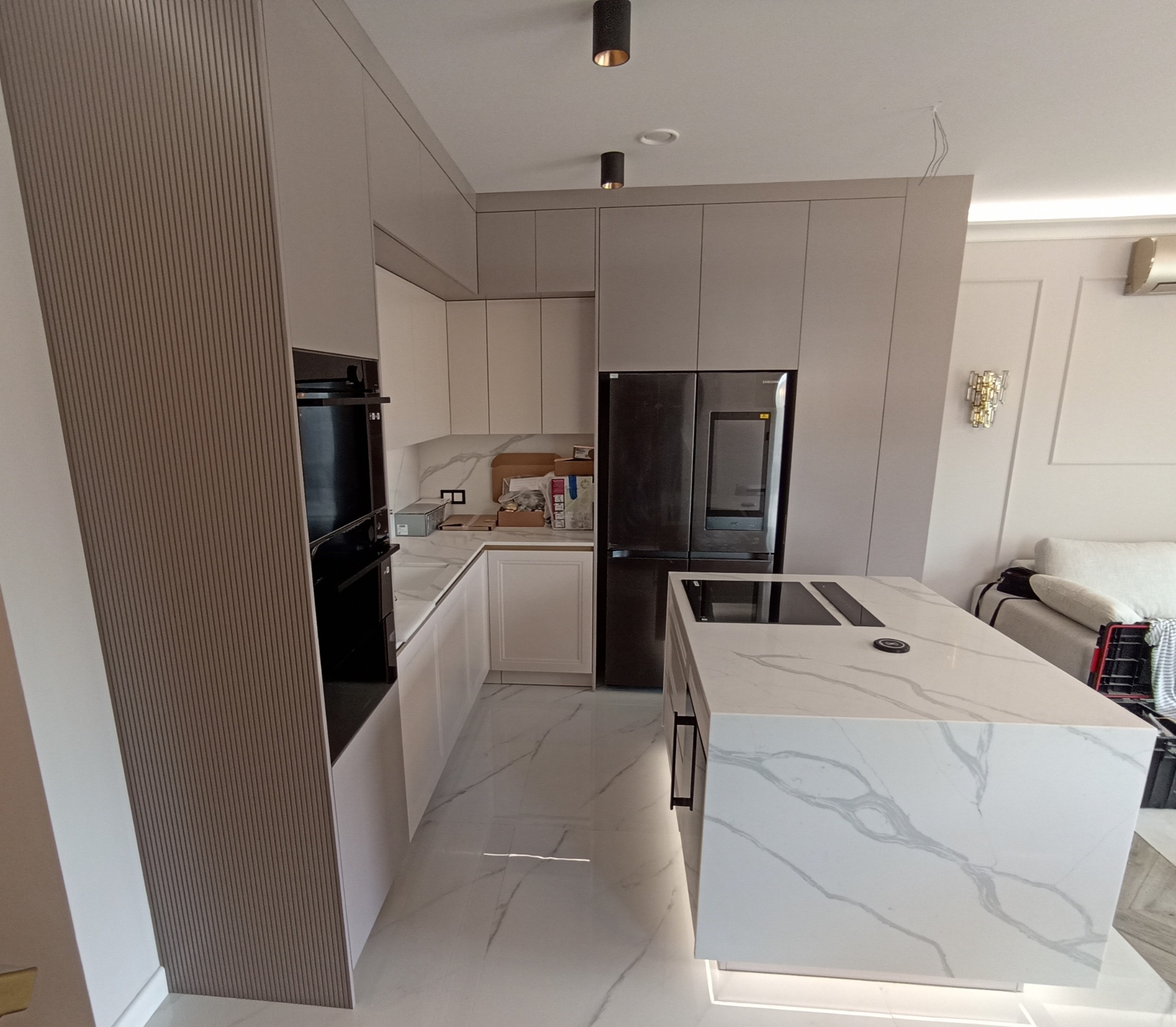 Nowoczesna kuchnia na wymiar w odcieniach szarości z wyspą o marmurowym blacie i podświetleniem LED. Widoczna zabudowa piekarnika i lodówka.