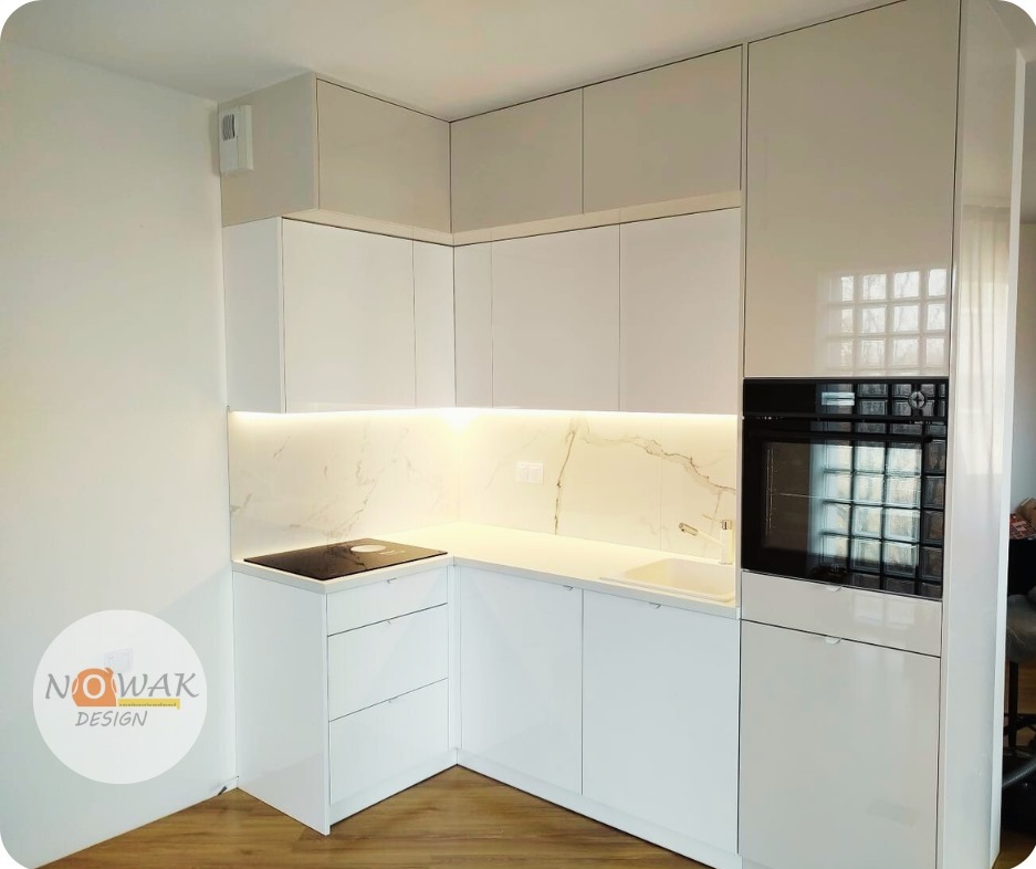 Biała, nowoczesna kuchnia z marmurowym blatem i oświetleniem LED. Widoczna płyta indukcyjna i piekarnik w zabudowie. Minimalistyczny design z połyskującymi frontami.
