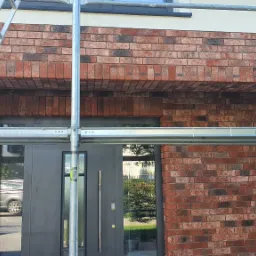 Fragment elewacji domu z czerwonej cegły klinkierowej, widoczne rusztowanie i czarne drzwi wejściowe z odbijającym się w szybie samochodem.