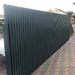 Nowoczesne, pionowe panele ogrodzeniowe z metalu w kolorze grafitowym, zamontowane na posesji z brukowaną nawierzchnią.
