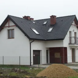 Dwukondygnacyjny dom jednorodzinny z ciemną dachówką, białymi ścianami i ceglanymi kominami, widoczny zza ogrodzenia z siatki.