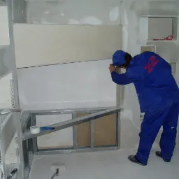 Pracownik w niebieskim kombinezonie szpachluje łączenie płyt kartonowo-gipsowych na ścianie w pomieszczeniu w trakcie prac wykończeniowych, widoczna metalowa konstrukcja pod płytami, taśma malarska...