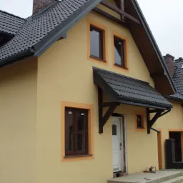 Nowo otynkowany dom jednorodzinny z ciemną dachówką, widoczne fragmenty rusztowania i resztki materiałów budowlanych wokół wejścia.