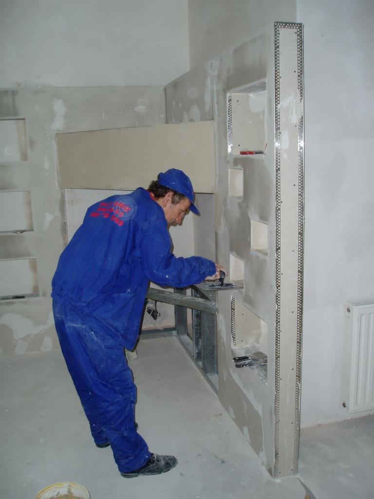 Pracownik w niebieskim kombinezonie szpachluje ścianę z płyty kartonowo-gipsowej z wnękami, widoczna metalowa konstrukcja, w tle surowe ściany w trakcie wykończenia i grzejnik.