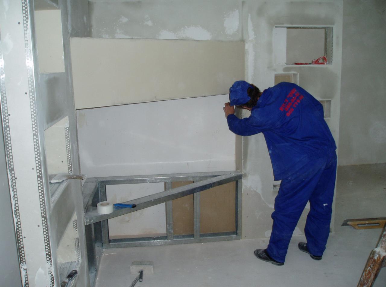 Pracownik w niebieskim kombinezonie szpachluje łączenie płyt kartonowo-gipsowych na ścianie w pomieszczeniu w trakcie prac wykończeniowych, widoczna metalowa konstrukcja pod płytami, taśma malarska...