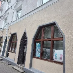 Elewacja kamienicy z oknami w kształcie łuku, widok z chodnika. Na elewacji widoczna rynna spustowa. Uwagę zwracają nietypowe okna i kolorystyka budynku.