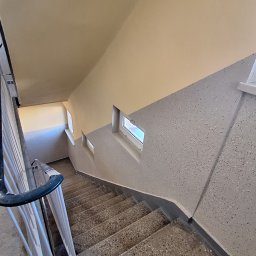 F.H.U MIESZBUD Grzegorz Mieszalski - Wnętrze klatki schodowej z widokiem na schody z lastryko, pomalowane ściany w odcieniach szarości i beżu oraz małe okna doświetlające przestrzeń.