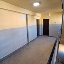 F.H.U MIESZBUD Grzegorz Mieszalski - Korytarz w bloku po odświeżeniu: ściany pomalowane na dwa kolory, żółty u góry i szary na dole. Widoczne drzwi do mieszkań i schody z metalową barierką.