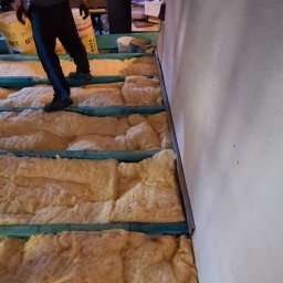 F.H.U MIESZBUD Grzegorz Mieszalski - Prace ociepleniowe na poddaszu: wełna mineralna ułożona między legarami, widoczna część sylwetki pracownika. Etap izolacji termicznej poddasza w trakcie realizacji.