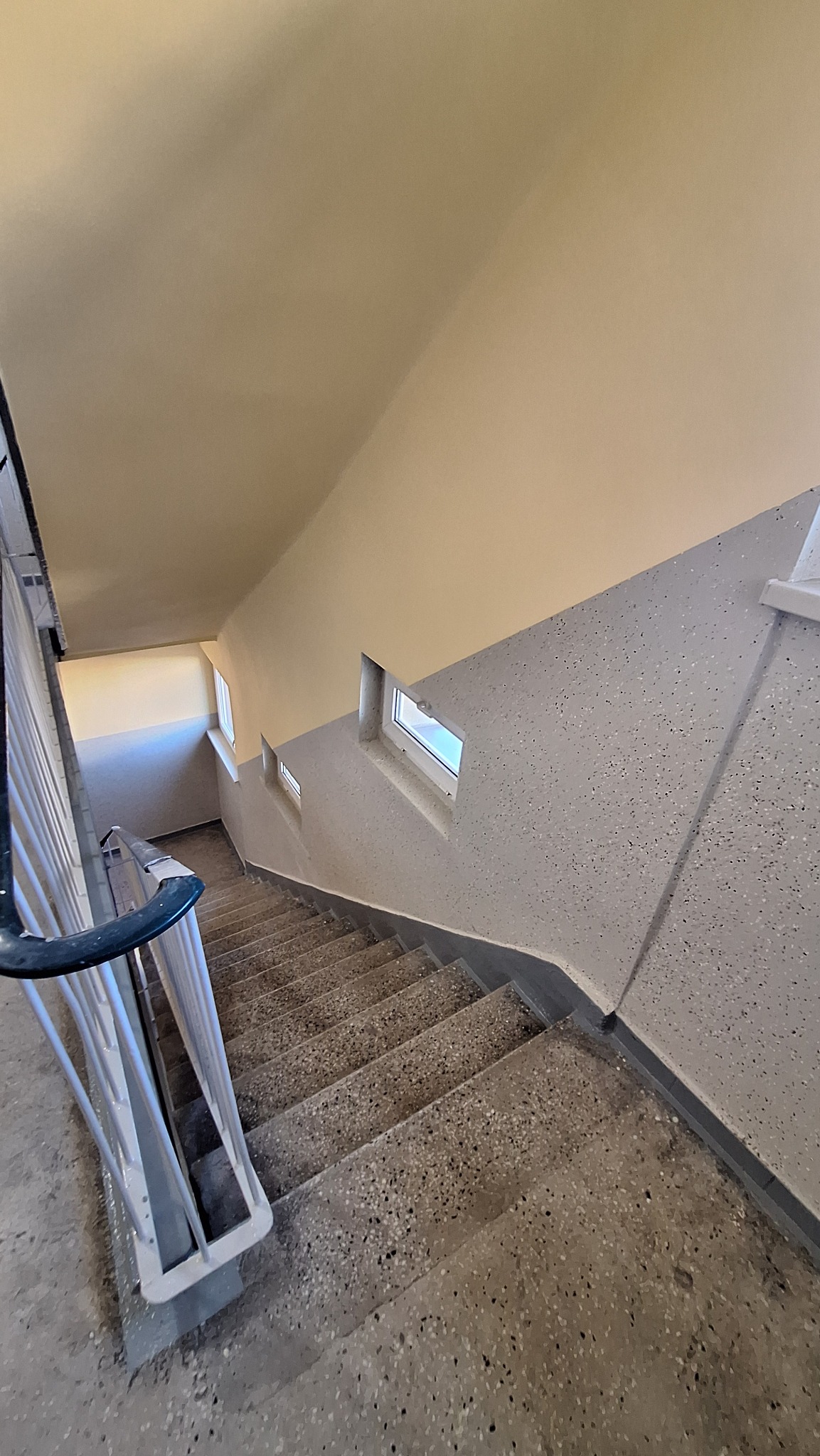 Wnętrze klatki schodowej z widokiem na schody z lastryko, pomalowane ściany w odcieniach szarości i beżu oraz małe okna doświetlające przestrzeń.
