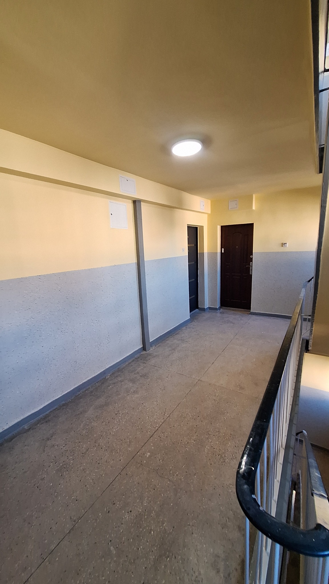 Korytarz w bloku po odświeżeniu: ściany pomalowane na dwa kolory, żółty u góry i szary na dole. Widoczne drzwi do mieszkań i schody z metalową barierką.