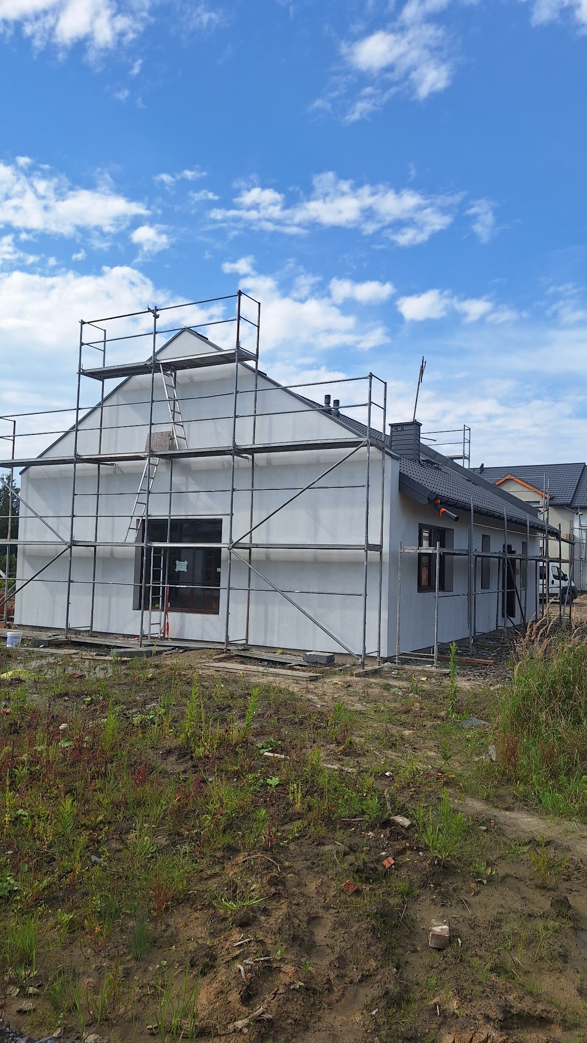 Dom w trakcie tynkowania elewacji, otoczony rusztowaniem. Widoczne fragmenty elewacji z szarym tynkiem. Niebo z chmurami w tle.