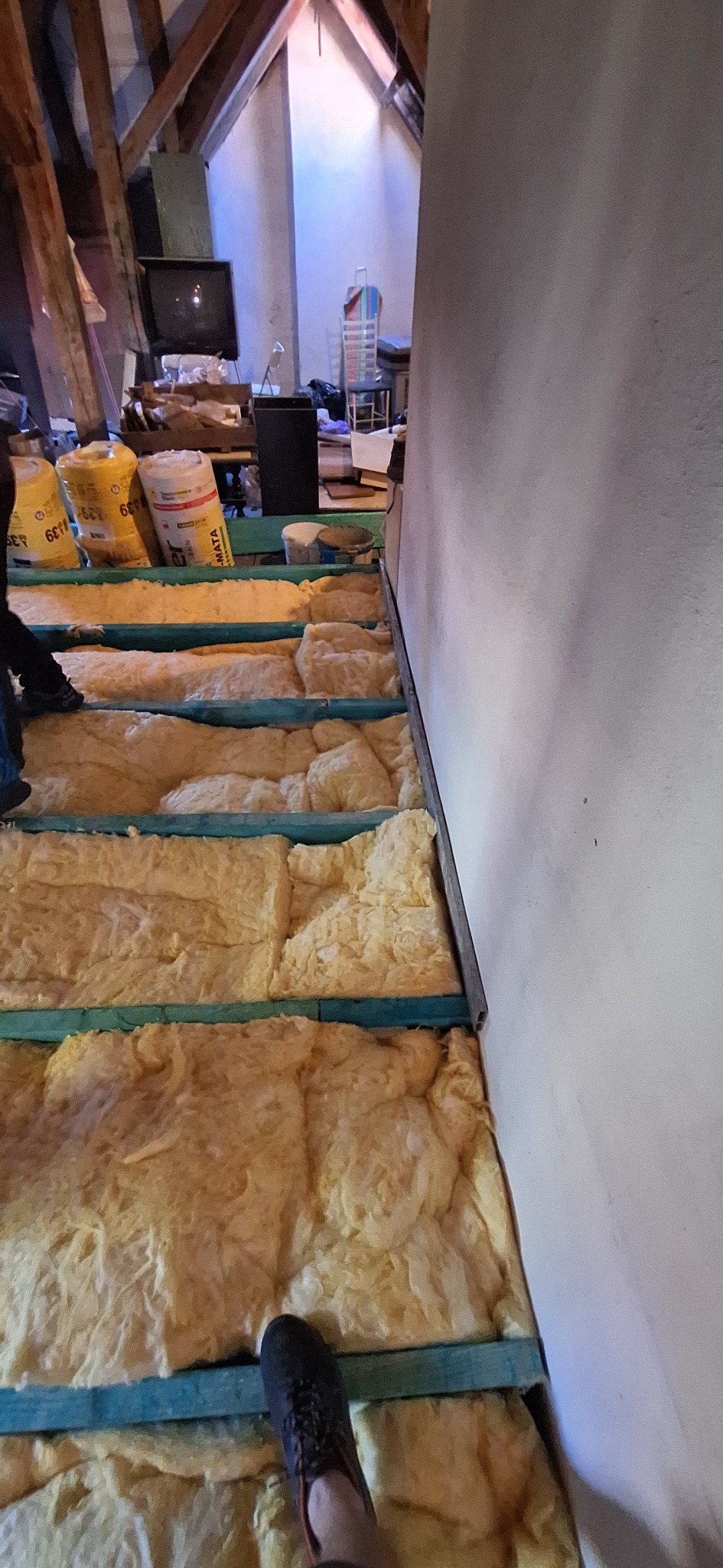 Ocieplenie poddasza wełną mineralną, widoczne drewniane legary i worki z materiałem izolacyjnym. Prace izolacyjne w trakcie realizacji na strychu.