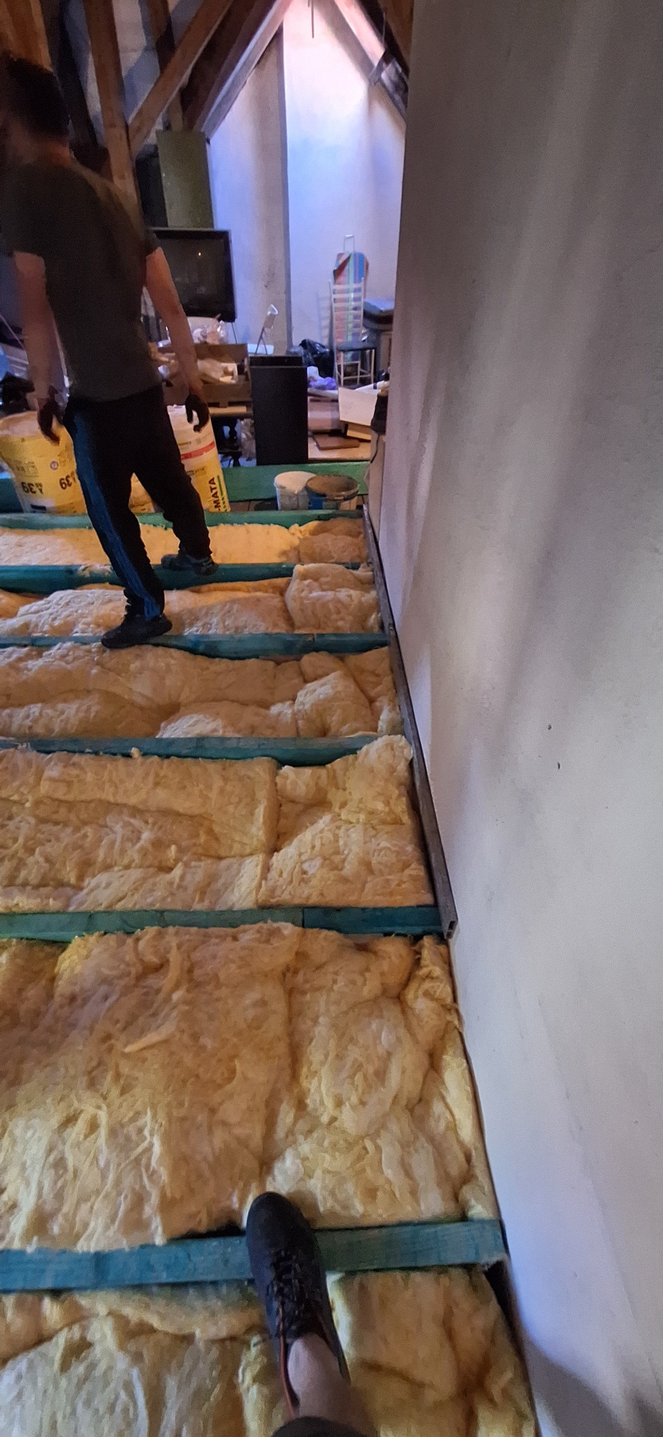 Prace ociepleniowe na poddaszu: wełna mineralna ułożona między legarami, widoczna część sylwetki pracownika. Etap izolacji termicznej poddasza w trakcie realizacji.