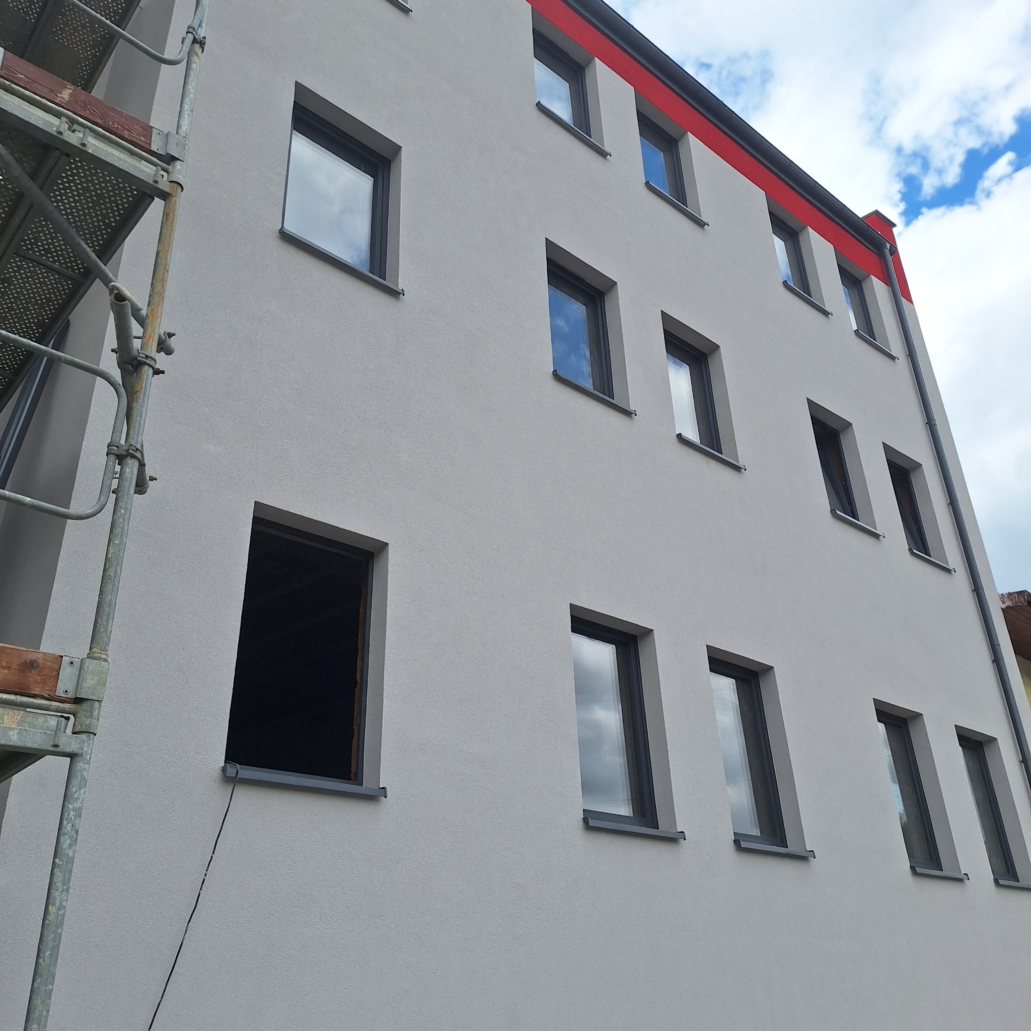 Szara elewacja budynku z czerwonym akcentem, widoczne okna z odbiciami nieba i fragment rusztowania z lewej strony. Jedno okno bez ramy.