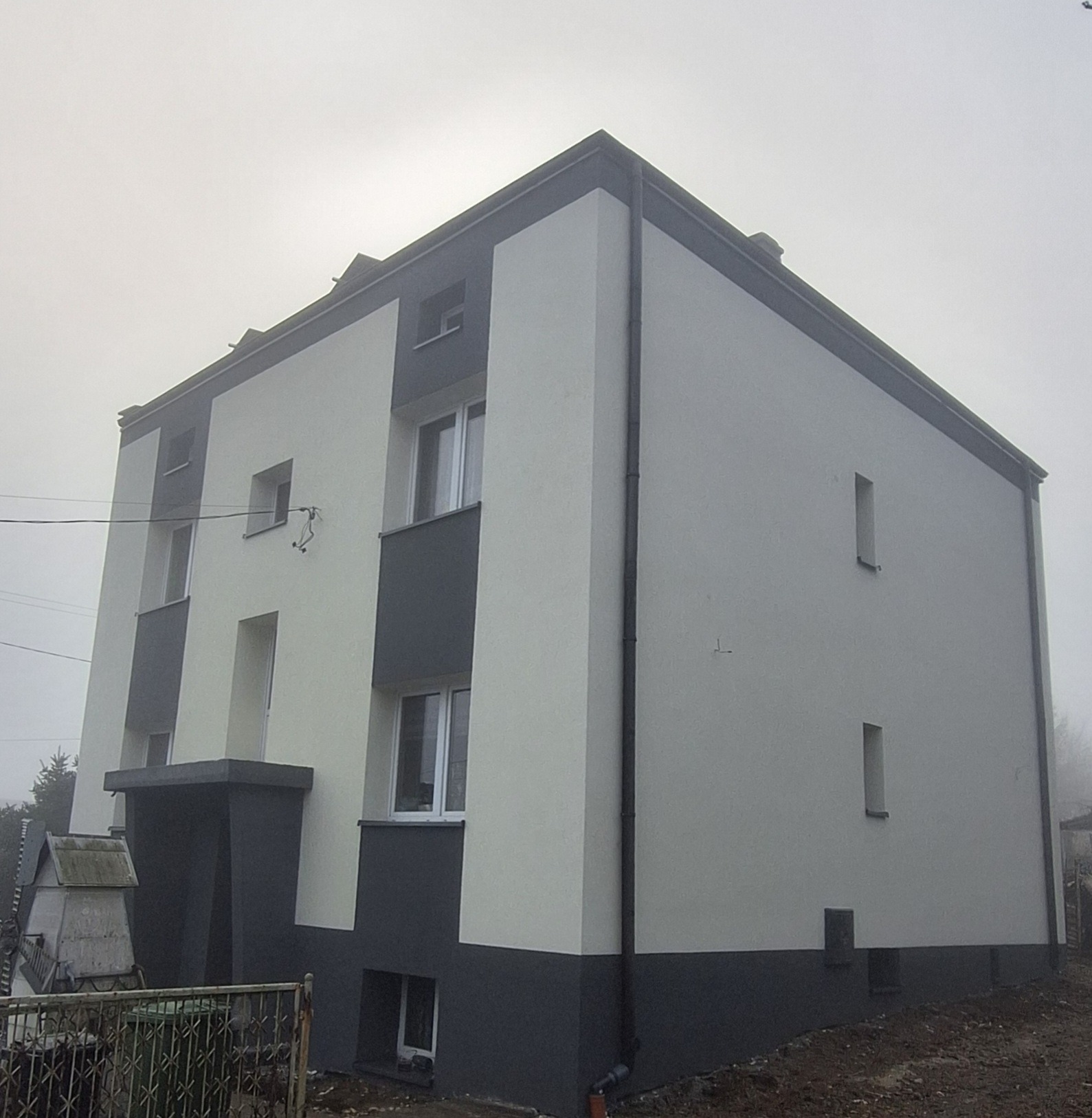 Dwukolorowa elewacja domu w mglisty dzień. Jasnoszary tynk kontrastuje z ciemnoszarymi elementami. Widoczne okna i rynny. Nowoczesny design.