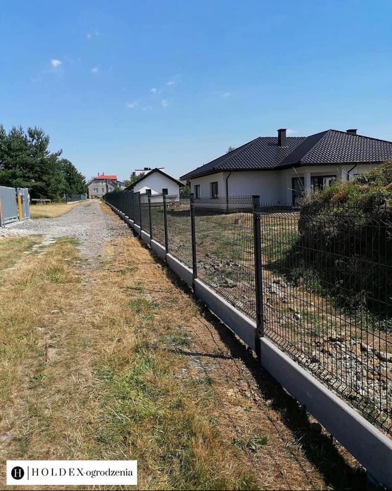 Długa, czarna siatka ogrodzeniowa na betonowej podmurówce, oddzielająca posesję z domem od polnej drogi z widocznymi śladami budowy.
