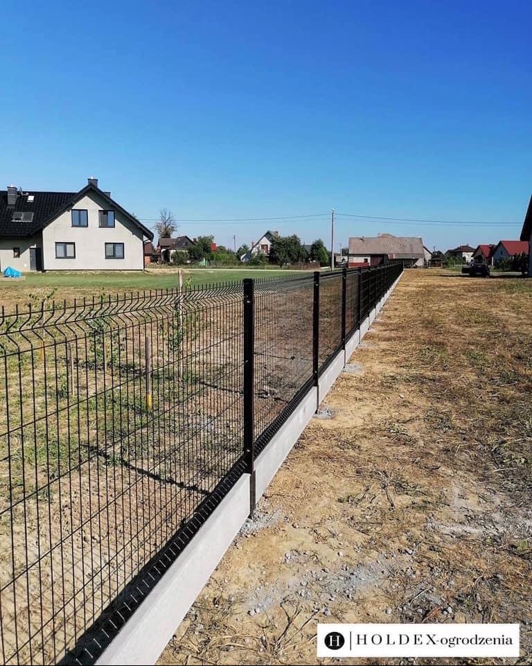 Nowoczesne ogrodzenie panelowe z podmurówką betonową, widok wzdłuż linii ogrodzenia w słoneczny dzień, domy w tle.