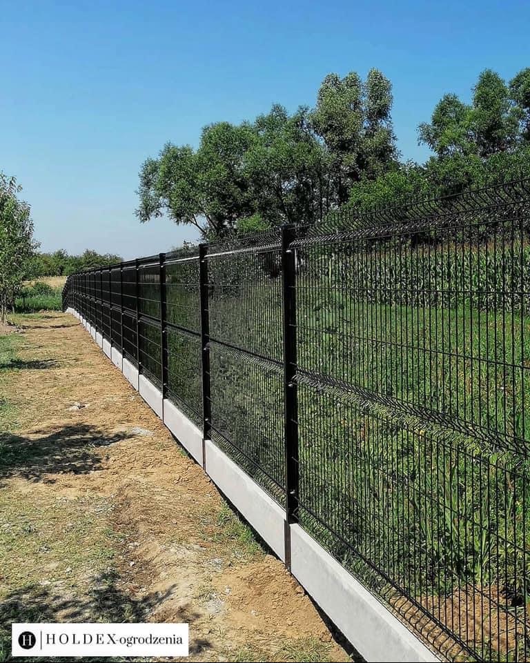 Długie, czarne ogrodzenie panelowe z podmurówką betonową, chroniące posesję z bujną roślinnością w słoneczny dzień.