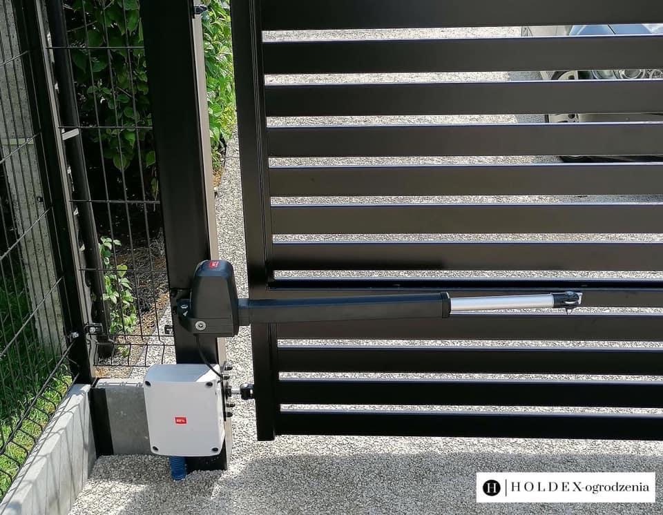 Automatyczna brama przesuwna z czarnych, poziomych profili, widoczny siłownik i centrala sterująca, na żwirowym podłożu, w tle zielona roślinność.