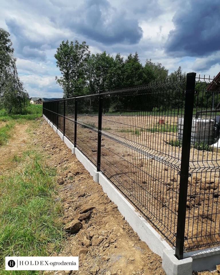 Długie, czarne ogrodzenie panelowe z siatki zgrzewanej, osadzone na betonowej podmurówce, widok z zewnątrz posesji, w tle drzewa i pochmurne niebo.