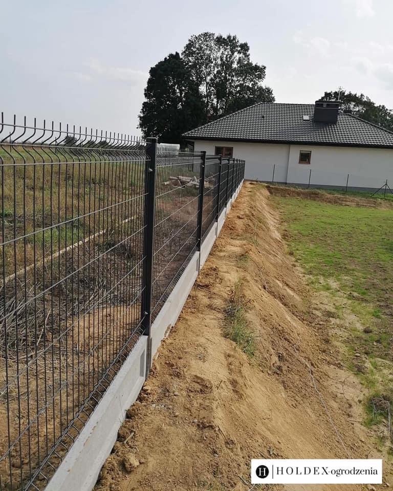 Nowoczesne ogrodzenie panelowe z podmurówką betonową, widok z boku, teren wokół jeszcze niezagospodarowany, w tle dom jednorodzinny z ciemną dachówką.